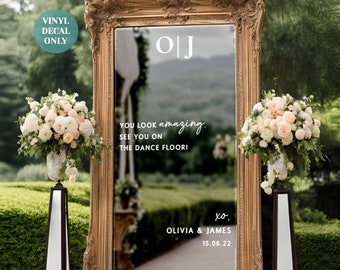 May include: A gold-framed mirror with a white vinyl decal that says "OJ" and "You look amazing. See you on the dance floor! xo. Olivia & James 15.06.22". The mirror is set up outdoors with floral arrangements on either side.