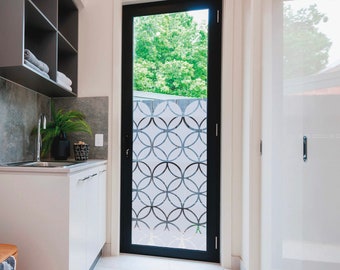 May include: A white door with a black frame and a geometric pattern etched into the glass. The door is open and you can see green trees through the glass.