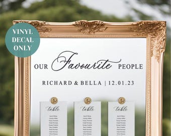 May include: A gold framed mirror with a white vinyl decal that reads "Our Favourite People Richard & Bella | 12.01.23".  The mirror is decorated with six white cards with gold accents that read "Table" and list names.