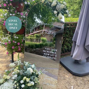 May include: A large, arched mirror with white text that reads "Welcome to Olga & Thomas Wedding". The mirror reflects a garden scene and is decorated with white flowers. Additional text says "You look incredible. Now let's celebrate!" and the date.