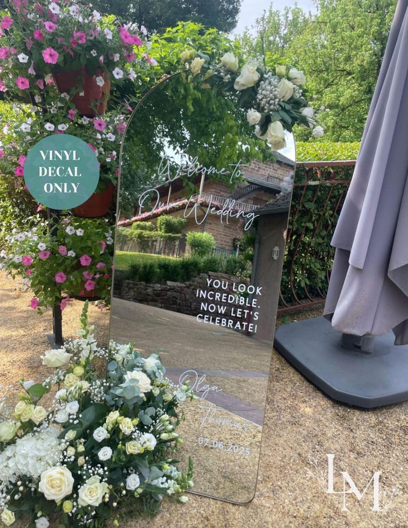 May include: A large, arched mirror with white text that reads "Welcome to Olga & Thomas Wedding". The mirror reflects a garden scene and is decorated with white flowers. Additional text says "You look incredible. Now let's celebrate!" and the date.