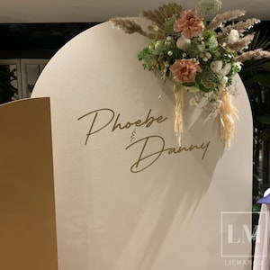 K&ouml;nnte beinhalten: Ein wei&szlig;er, halbkreisf&ouml;rmiger Hintergrund mit einer goldenen Aufschrift, die "Phoebe & Danny" lautet. Der Hintergrund ist mit einem Strau&szlig; getrockneter Blumen und Gr&auml;ser verziert.