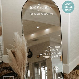 May include: An arched floor mirror featuring white vinyl decal text that reads welcome TO OUR WEDDING, YOU LOOK INCREDIBLE. WE'RE SO GLAD YOU'RE HERE! and xo J | L. The mirror has a thin black frame and is displayed with a vase of dried pampas grass.