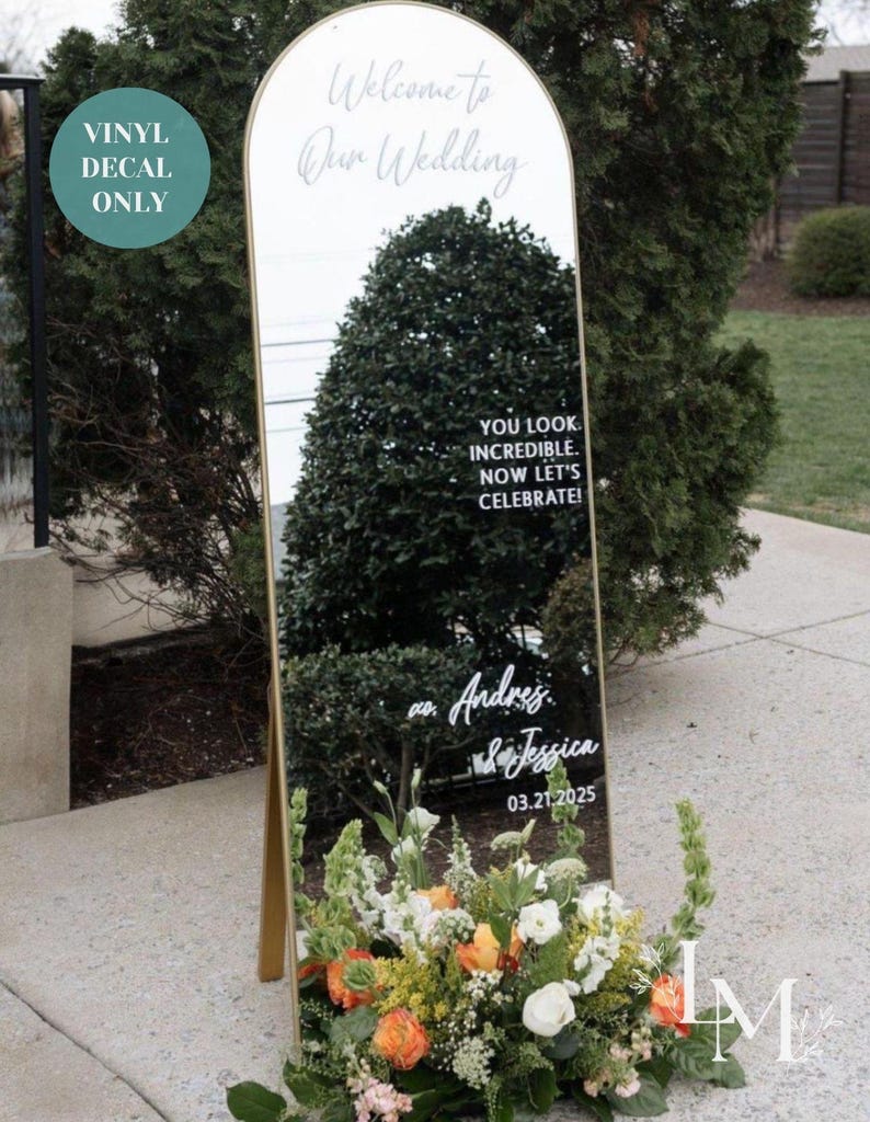 May include: A tall, arched mirror with a gold frame serves as a wedding welcome sign. The mirror reflects the surroundings and features elegant white script that reads "Welcome to Our Wedding," along with the names Andres & Jessica and the date 03.21.2025. A floral arrangement is at the base.