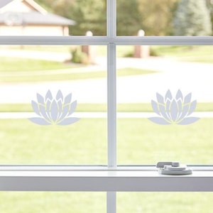 May include: Two white lotus flower decals are applied to a window pane. The decals are in the centre of each pane. The window is white and has a white frame. The window is open and there is a green lawn visible through the window.