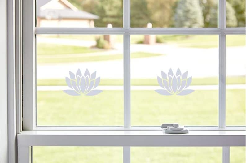 May include: Two white lotus flower decals are applied to a window pane. The decals are in the centre of each pane. The window is white and has a white frame. The window is open and there is a green lawn visible through the window.