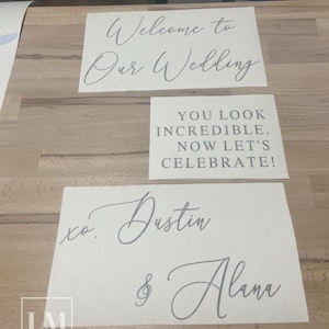 Op de afbeelding: Drie witte rechthoekige vinylstickers met grijze letters. De bovenste sticker bevat de tekst Welcome to Our Wedding in cursief schrift. De middelste sticker bevat YOU LOOK INCREDIBLE. NOW LET'S CELEBRATE! in blokletters. De onderste sticker bevat xo. Dustin & Alana.