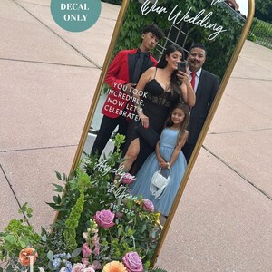May include: A gold-framed mirror with the text "Welcome to Our Wedding" and "Angelique & Oscar 8th, 2025". The mirror reflects a family. A floral arrangement with pink, purple, and orange flowers is at the base. A teal circle reads "VINYL DECAL ONLY".