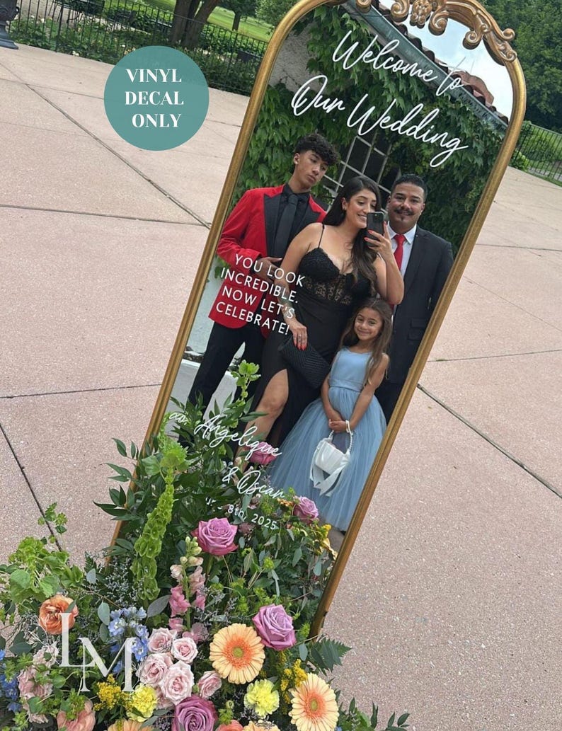 May include: A gold-framed mirror with the text "Welcome to Our Wedding" and "Angelique & Oscar 8th, 2025". The mirror reflects a family. A floral arrangement with pink, purple, and orange flowers is at the base. A teal circle reads "VINYL DECAL ONLY".