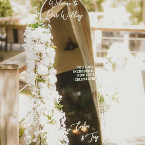 Op de afbeelding: Een boogvormig spiegel-bruiloftsbord met een gouden lijst, versierd met een verticaal arrangement van witte bloemen. De tekst luidt: Welcome to Our Wedding, YOU LOOK INCREDIBLE. NOW LET'S CELEBRATE!, Evelyn & Tony, en de datum 30-08-2025.