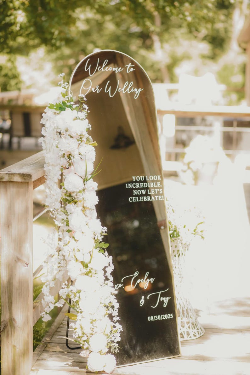 Op de afbeelding: Een boogvormig spiegel-bruiloftsbord met een gouden lijst, versierd met een verticaal arrangement van witte bloemen. De tekst luidt: Welcome to Our Wedding, YOU LOOK INCREDIBLE. NOW LET'S CELEBRATE!, Evelyn & Tony, en de datum 30-08-2025.