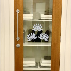 May include: A wooden door with a glass panel featuring a white lotus flower design. The glass panel reveals a white shelf with folded towels and a basket.