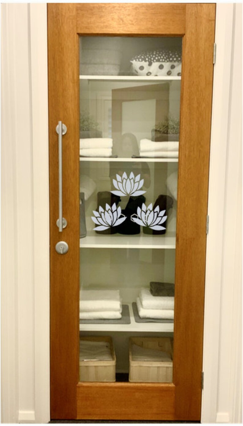 May include: A wooden door with a glass panel featuring a white lotus flower design. The glass panel reveals a white shelf with folded towels and a basket.