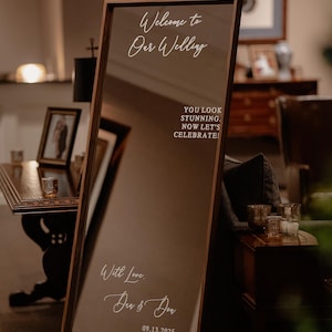 Op de afbeelding: Een hoge rechthoekige spiegel met een donkerhouten frame, voorzien van witte tekst: Welcome to Our Wedding, YOU LOOK STUNNING. NOW LET'S CELEBRATE!, With Love, Den & Don, en 09.13.2025. De spiegel staat op een witte metalen standaard.