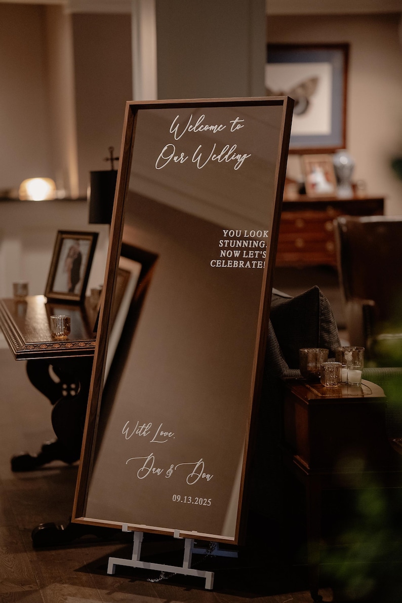 Op de afbeelding: Een hoge rechthoekige spiegel met een donkerhouten frame, voorzien van witte tekst: Welcome to Our Wedding, YOU LOOK STUNNING. NOW LET'S CELEBRATE!, With Love, Den & Don, en 09.13.2025. De spiegel staat op een witte metalen standaard.