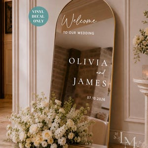 May include: An arched floor mirror with a gold frame featuring white vinyl decal text that reads Welcome TO OUR WEDDING OLIVIA and JAMES 07.10.2026. A teal circular badge in the top left corner states VINYL DECAL ONLY. The mirror reflects a room interior.