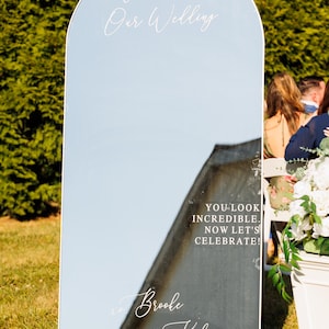 Op de afbeelding: Een boogvormig spiegel-bruiloftsbord met witte bloemstukken aan de boven- en onderkant. De tekst luidt: Welcome to Our Wedding, YOU LOOK INCREDIBLE. NOW LET'S CELEBRATE!, Brooke & Kyle, en 09.06.2025. De spiegel reflecteert de omliggende buitenomgeving.