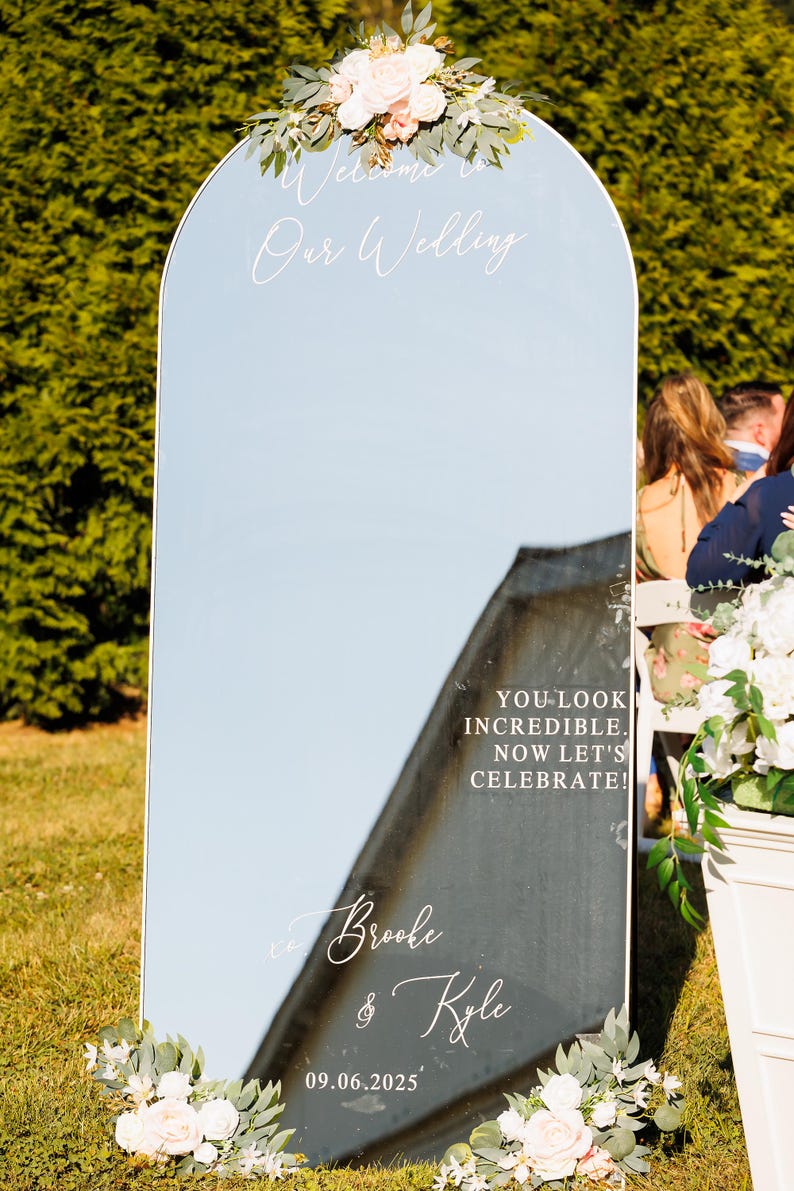Op de afbeelding: Een boogvormig spiegel-bruiloftsbord met witte bloemstukken aan de boven- en onderkant. De tekst luidt: Welcome to Our Wedding, YOU LOOK INCREDIBLE. NOW LET'S CELEBRATE!, Brooke & Kyle, en 09.06.2025. De spiegel reflecteert de omliggende buitenomgeving.
