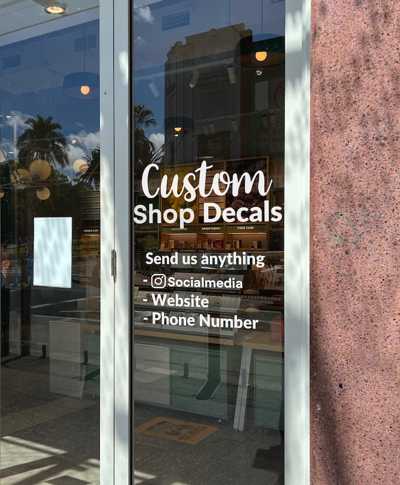 K&ouml;nnte beinhalten: Ein wei&szlig;es Schaufenster mit einem Schild, auf dem "Custom Shop Decals" steht. Das Schild enth&auml;lt auch den Text "Send us anything" und eine Liste von M&ouml;glichkeiten, den Shop zu kontaktieren: "Social Media", "Website" und "Phone Number".