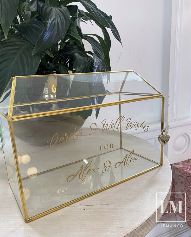 K&ouml;nnte beinhalten: Eine Kartenbox aus Glas und Gold mit einem herzf&ouml;rmigen Schloss. Die Box tr&auml;gt ein Schild mit der Aufschrift "All Cards & Well Wishes for Alex & Alex".