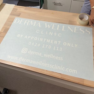 K&ouml;nnte beinhalten: Ein wei&szlig;es Vinyl-Schild mit dem Text "Derma Wellness Clinic" in einer modernen Schriftart. Das Schild enth&auml;lt auch den Text "Nur nach Vereinbarung", eine Telefonnummer, einen Social-Media-Handle und eine Website-Adresse.