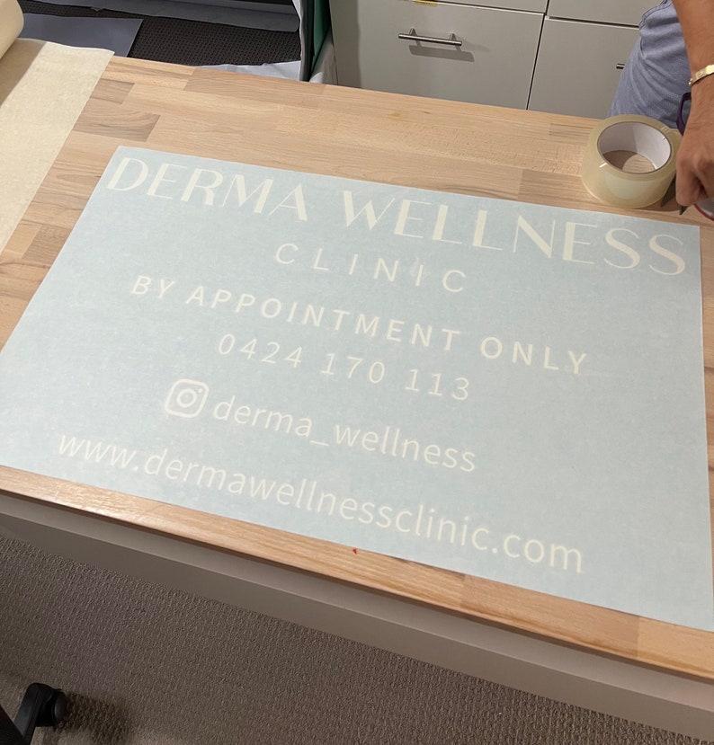 K&ouml;nnte beinhalten: Ein wei&szlig;es Vinyl-Schild mit dem Text "Derma Wellness Clinic" in einer modernen Schriftart. Das Schild enth&auml;lt auch den Text "Nur nach Vereinbarung", eine Telefonnummer, einen Social-Media-Handle und eine Website-Adresse.