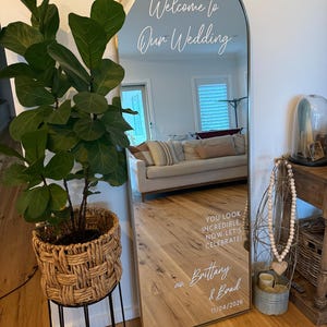 Op de afbeelding: Een boogvormige vloerspiegel met een dunne gouden lijst en witte vinyltekst met de tekst Welcome to Our Wedding, YOU LOOK INCREDIBLE. NOW LET'S CELEBRATE!, Brittany & Brad en 11/04/2026. De spiegel reflecteert een woonkamerinterieur.