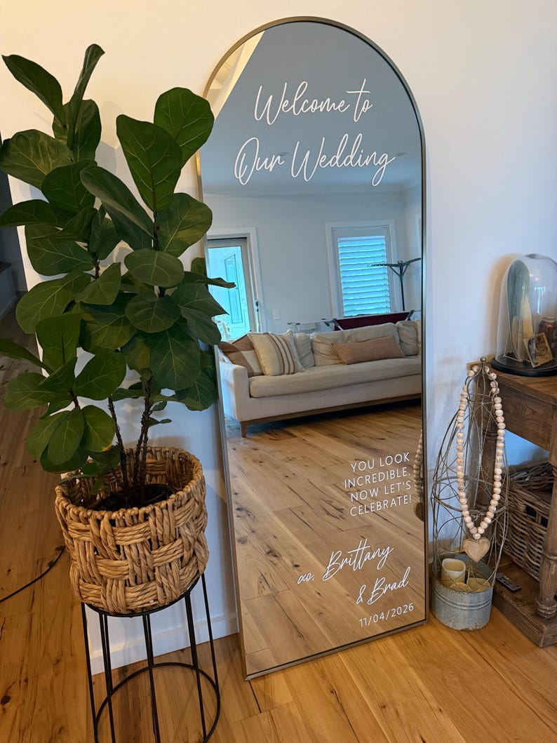 Op de afbeelding: Een boogvormige vloerspiegel met een dunne gouden lijst en witte vinyltekst met de tekst Welcome to Our Wedding, YOU LOOK INCREDIBLE. NOW LET'S CELEBRATE!, Brittany & Brad en 11/04/2026. De spiegel reflecteert een woonkamerinterieur.