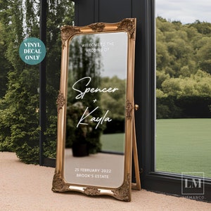May include: A gold-framed mirror with a white vinyl decal announcing a wedding. The decal reads "Welcome to the Wedding of Spencer + Kayla" and includes the date "25 February 2022" and the location "Brook's Estate".