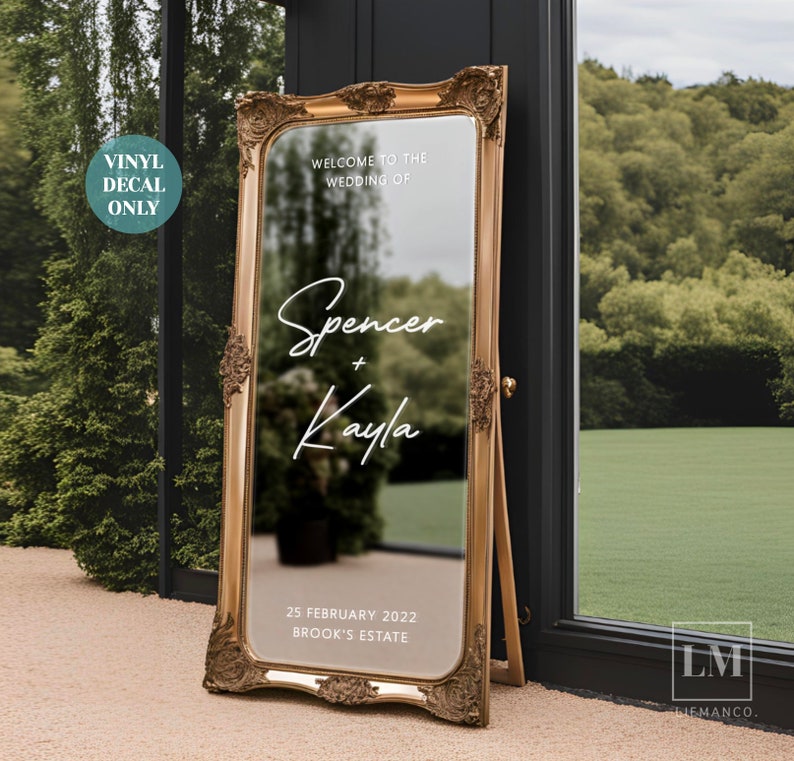 May include: A gold-framed mirror with a white vinyl decal announcing a wedding. The decal reads "Welcome to the Wedding of Spencer + Kayla" and includes the date "25 February 2022" and the location "Brook's Estate".