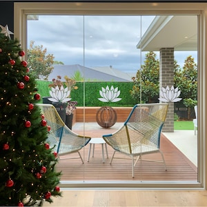 May include: A Christmas tree decorated with red baubles and white fairy lights is in the foreground. A sliding glass door leads to a patio with two chairs and a small table. The patio has a wooden deck and a green lawn. The glass door has white lotus flower decals.