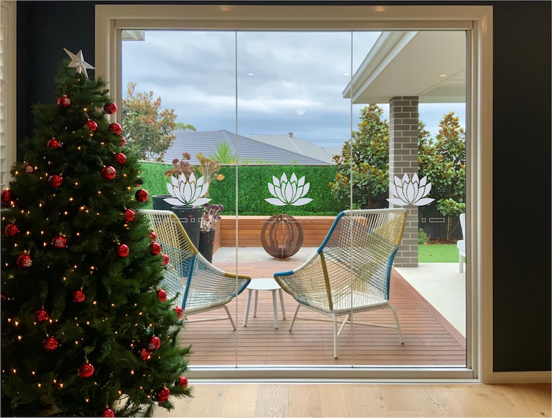 May include: A Christmas tree decorated with red baubles and white fairy lights is in the foreground. A sliding glass door leads to a patio with two chairs and a small table. The patio has a wooden deck and a green lawn. The glass door has white lotus flower decals.