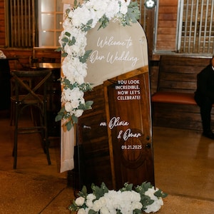 Op de afbeelding: Een boogvormig spiegel-bruiloftsbord met witte bloemstukken linksboven en onderaan. De tekst luidt: Welcome to Our Wedding, YOU LOOK INCREDIBLE. NOW LET'S CELEBRATE!, xo. Olivia & Omar, 09.21.2025. De spiegel reflecteert een houten deur.