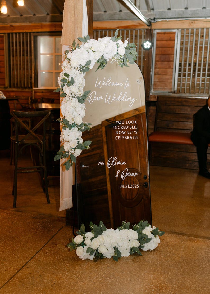 Op de afbeelding: Een boogvormig spiegel-bruiloftsbord met witte bloemstukken linksboven en onderaan. De tekst luidt: Welcome to Our Wedding, YOU LOOK INCREDIBLE. NOW LET'S CELEBRATE!, xo. Olivia & Omar, 09.21.2025. De spiegel reflecteert een houten deur.