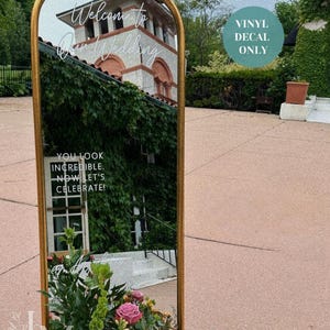 May include: A gold-framed mirror with ornate detailing at the top, reflecting a building covered in ivy. The mirror has the text "Welcome to Our Wedding" and "You look incredible. Now let's celebrate!" A floral arrangement with pink and orange flowers is at the base.