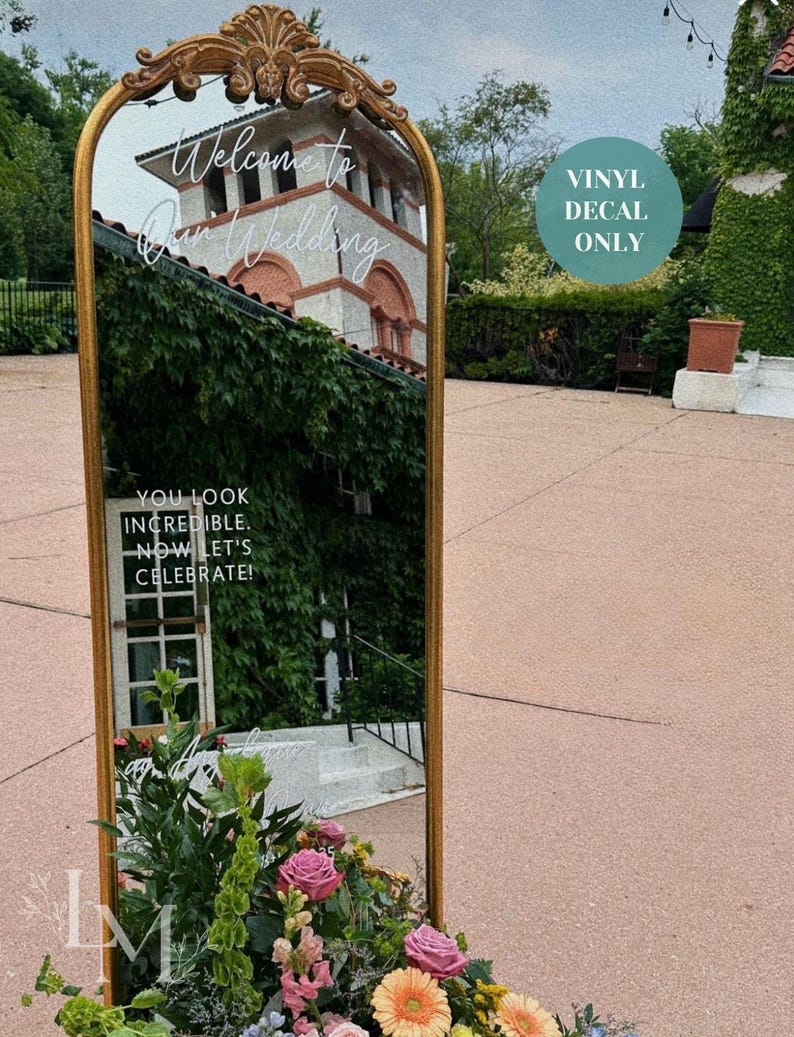 May include: A gold-framed mirror with ornate detailing at the top, reflecting a building covered in ivy. The mirror has the text "Welcome to Our Wedding" and "You look incredible. Now let's celebrate!" A floral arrangement with pink and orange flowers is at the base.
