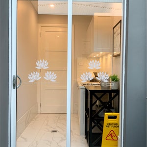 May include: A view through a glass door into a kitchen with white cabinets, a black metal shelf, and a yellow caution wet floor sign. The glass door has a white lotus flower design.