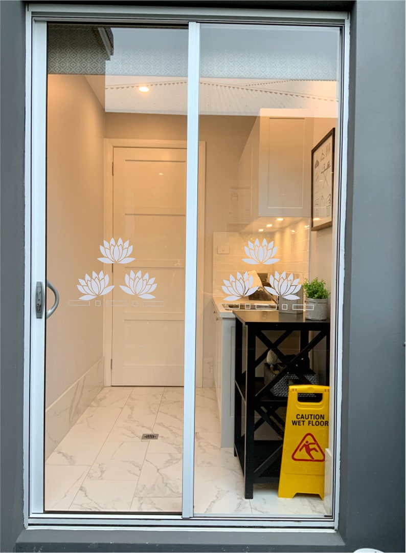 May include: A view through a glass door into a kitchen with white cabinets, a black metal shelf, and a yellow caution wet floor sign. The glass door has a white lotus flower design.