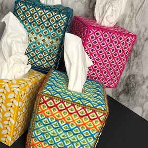 May include: A collection of decorative tissue box covers in various colors. The covers are square-shaped and feature intricate embroidered patterns in teal, pink, and rainbow hues. Each box has a white tissue emerging from the top. One box is yellow with a leaf pattern.