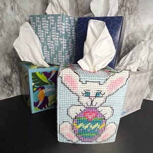 May include: A rectangular tissue box cover made of light blue plastic canvas with a cross-stitch design of a white bunny holding a colorful patterned Easter egg. The bunny has pink ears and blue eyes. It is displayed among several other tissue boxes.