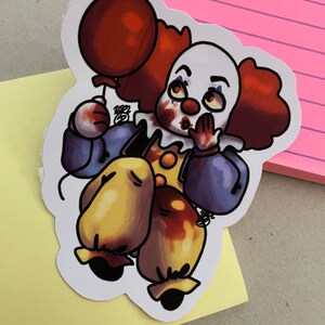 May include: A sticker of a cartoon clown with red hair, a white face, and a red balloon. The clown is wearing a blue and yellow costume and has a surprised expression. The sticker is on a yellow background.