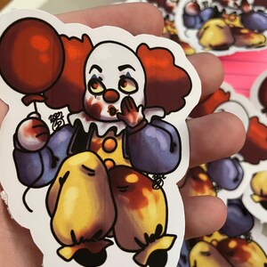 May include: A sticker of a cartoon clown with red hair, a white face, and a red balloon. The clown is wearing a blue and purple outfit with yellow accents. The clown has a red nose and is holding the balloon in its left hand. The clown is smiling and has a mischievous look on its face.