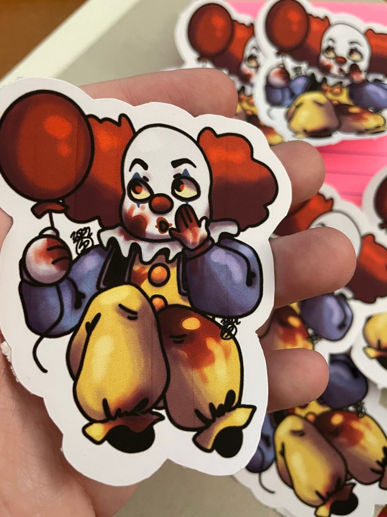 May include: A sticker of a cartoon clown with red hair, a white face, and a red balloon. The clown is wearing a blue and purple outfit with yellow accents. The clown has a red nose and is holding the balloon in its left hand. The clown is smiling and has a mischievous look on its face.