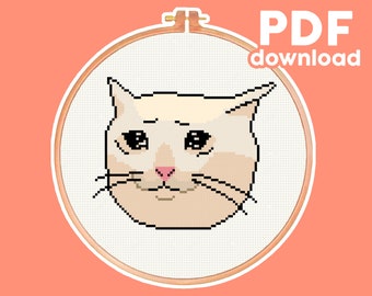 May include: Cross-stitch pattern of a white cat with a sad expression, in a wooden embroidery hoop. The cat has black eyes and a pink nose. The pattern is available as a PDF download.
