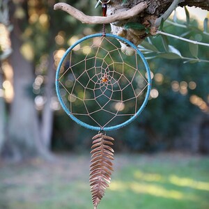 Puede incluir: Un atrapasueños turquesa con una pluma de cobre colgando de él. El atrapasueños está colgado de una rama en un jardín.