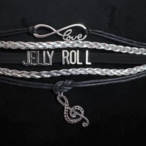May include: A collection of bracelets with black and silver cords. One bracelet has the word "love" within an infinity symbol. Another bracelet has the words "JELLY ROLL". A treble clef charm hangs from the bottom bracelet.