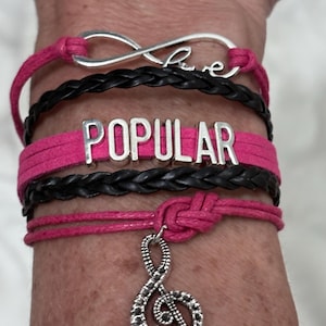 May include: A stack of bracelets in pink and black. One bracelet has the word "POPULAR" in silver letters. Another has the word "love" in cursive. A silver treble clef charm hangs from a pink bracelet.