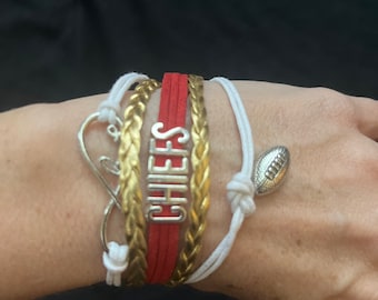 May include: A set of bracelets featuring a red braided bracelet with the word "CHIEFS" in silver lettering, a gold braided bracelet, a white braided bracelet with a silver football charm, and a white infinity symbol charm.