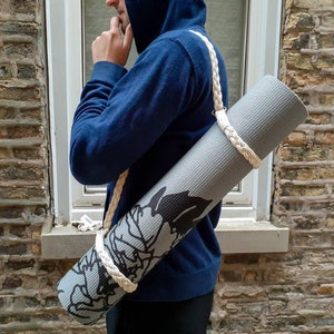 May include: A person carrying a grey yoga mat with a black and white floral design. The mat is secured with a white rope strap.