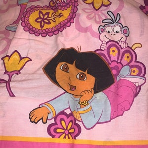Może przedstawiać: Różowa poszwa na pościel z Dorą poznawczynią, uśmiechniętą postacią z kreskówki. Poszwa ma kwiatowy i paisley wzór w odcieniach różu, żółci i fioletu. Widoczna jest również postać małpy.